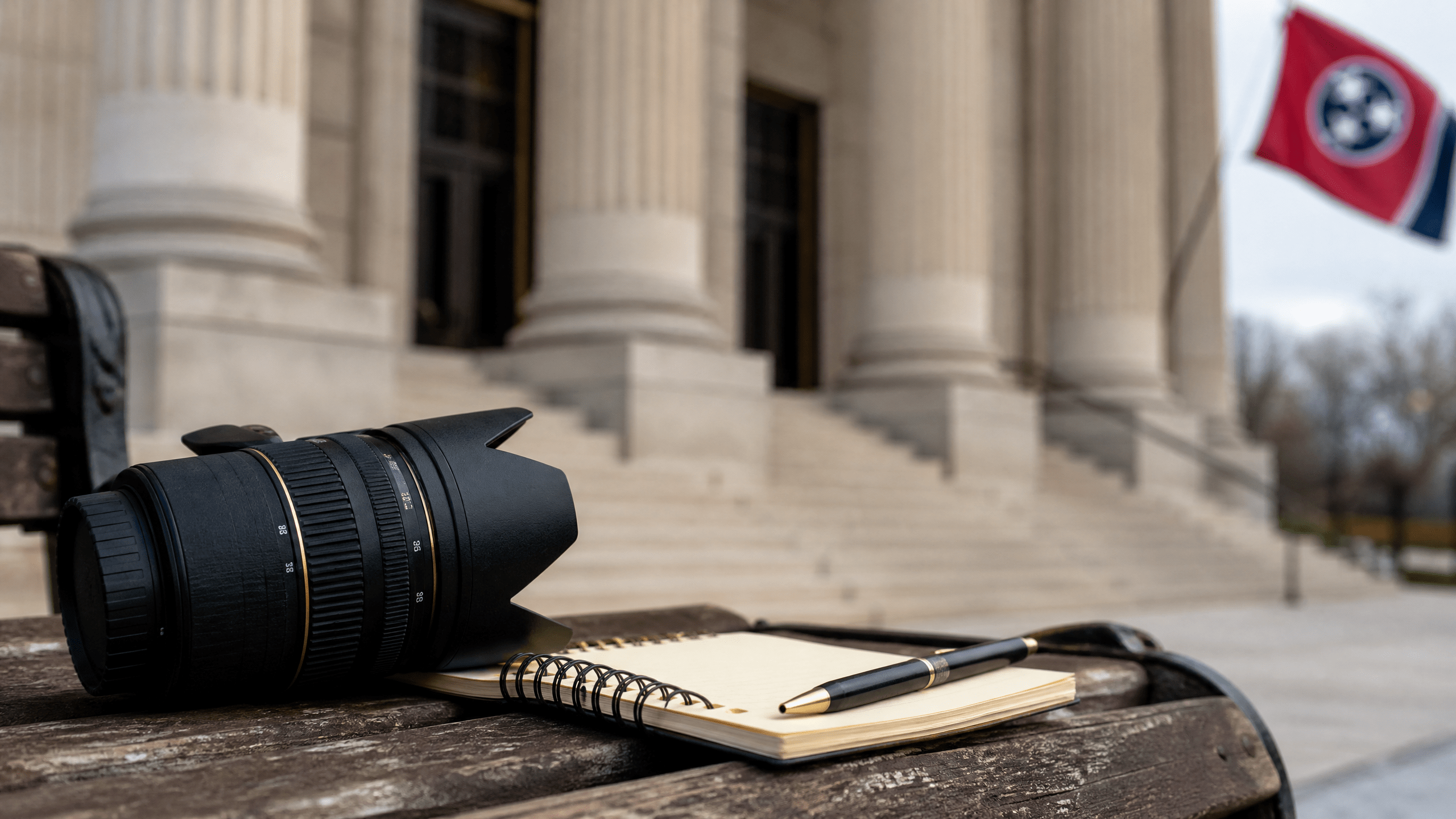 A camera lens and notebook rest on a wooden bench outside a courtroom, illustrating media access.